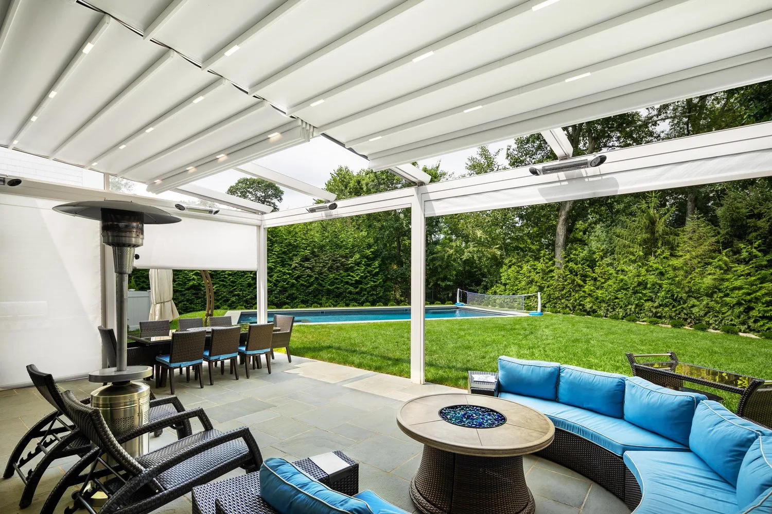 Wider view of the same white frame from the fire table, heater and dining set beneath the closed membrane, lawn and pool beyond — photograph