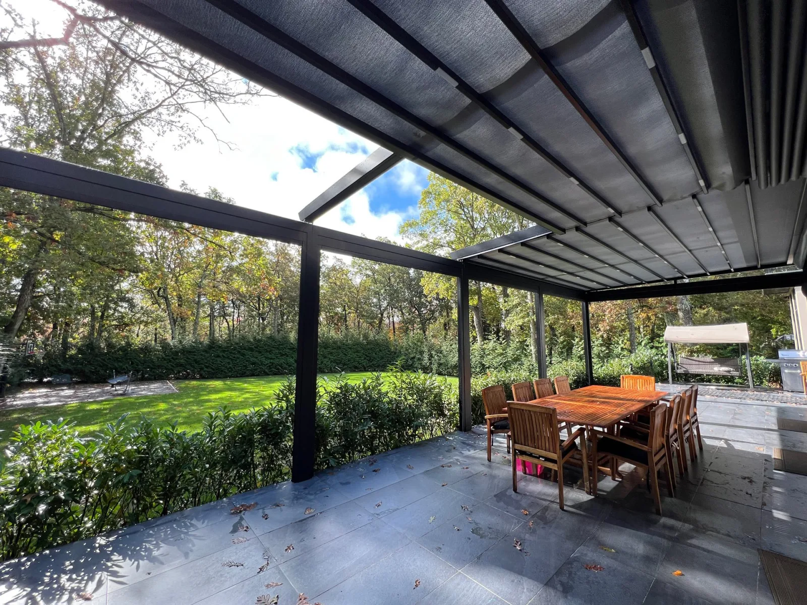 Dark-framed retractable fabric roof caught mid-run over a bluestone patio, open sky ahead of the front bar and taut fabric behind — photograph