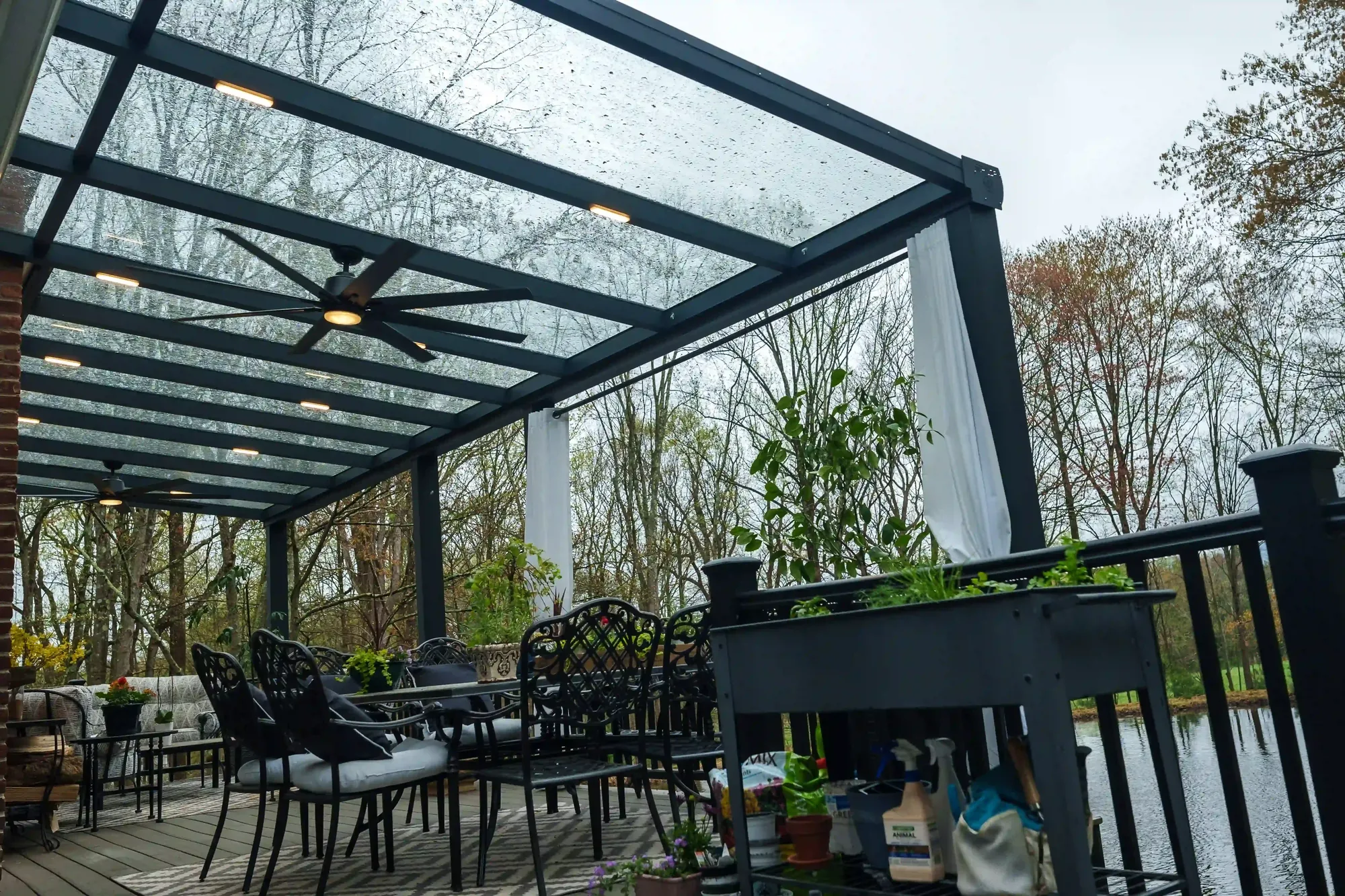 Rain beaded across a glass pergola roof over a waterside deck, the seating dry beneath — photograph