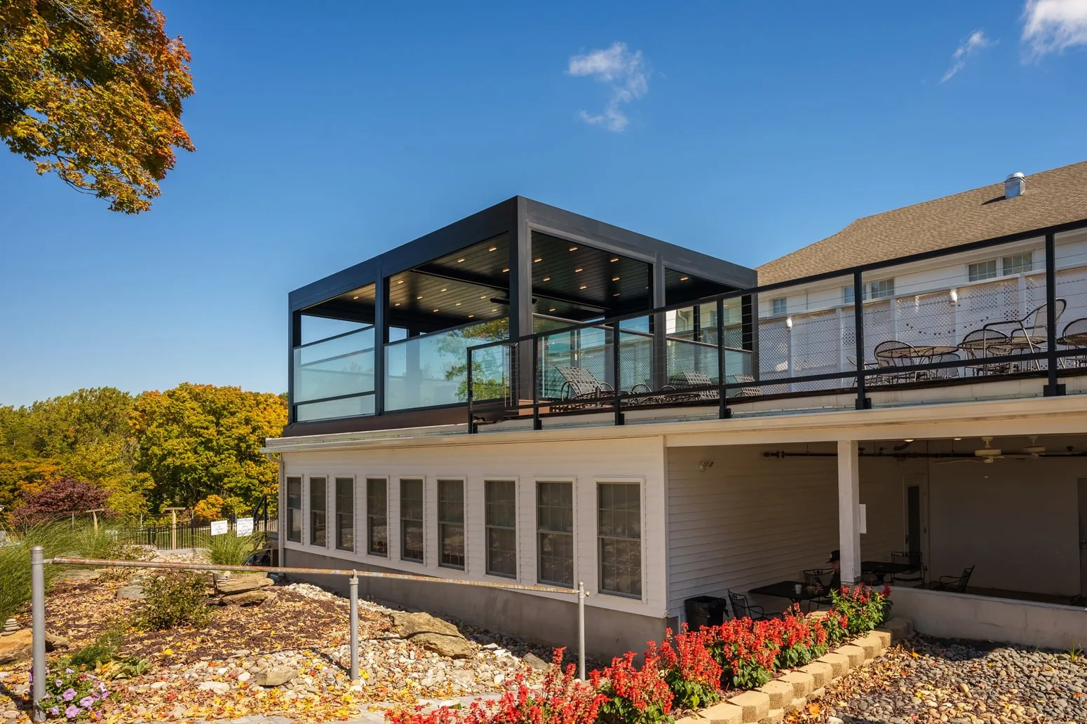 A dark-framed glass room raised on a deck above an autumn garden, glazed on every side with the panes closed — photograph