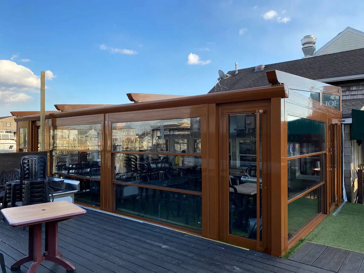 A wood-toned framed enclosure closing a deck, glazed corner to corner with tables and stacked chairs visible through the panes — photograph