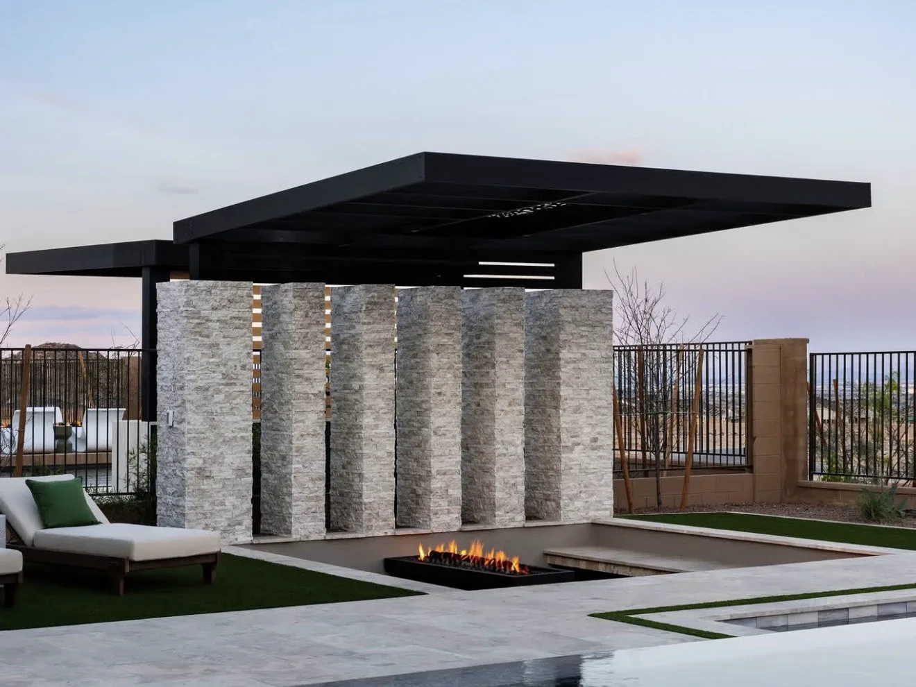 Hard top roof carried on a stacked stone wall with lit vertical slots, fire trough and pool below at dusk — photograph