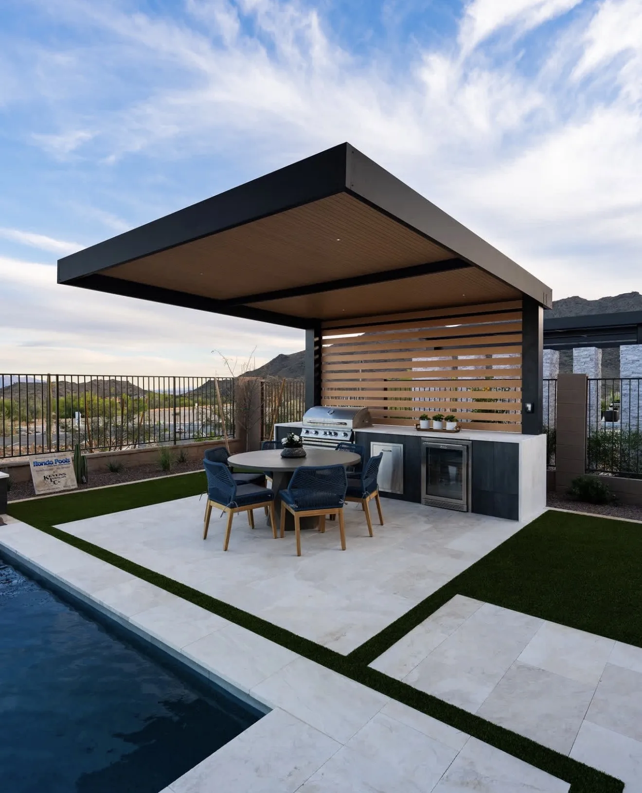 Hard top pergola with a warm timber soffit over an outdoor kitchen and round dining table, slatted screen behind and mountains beyond — photograph