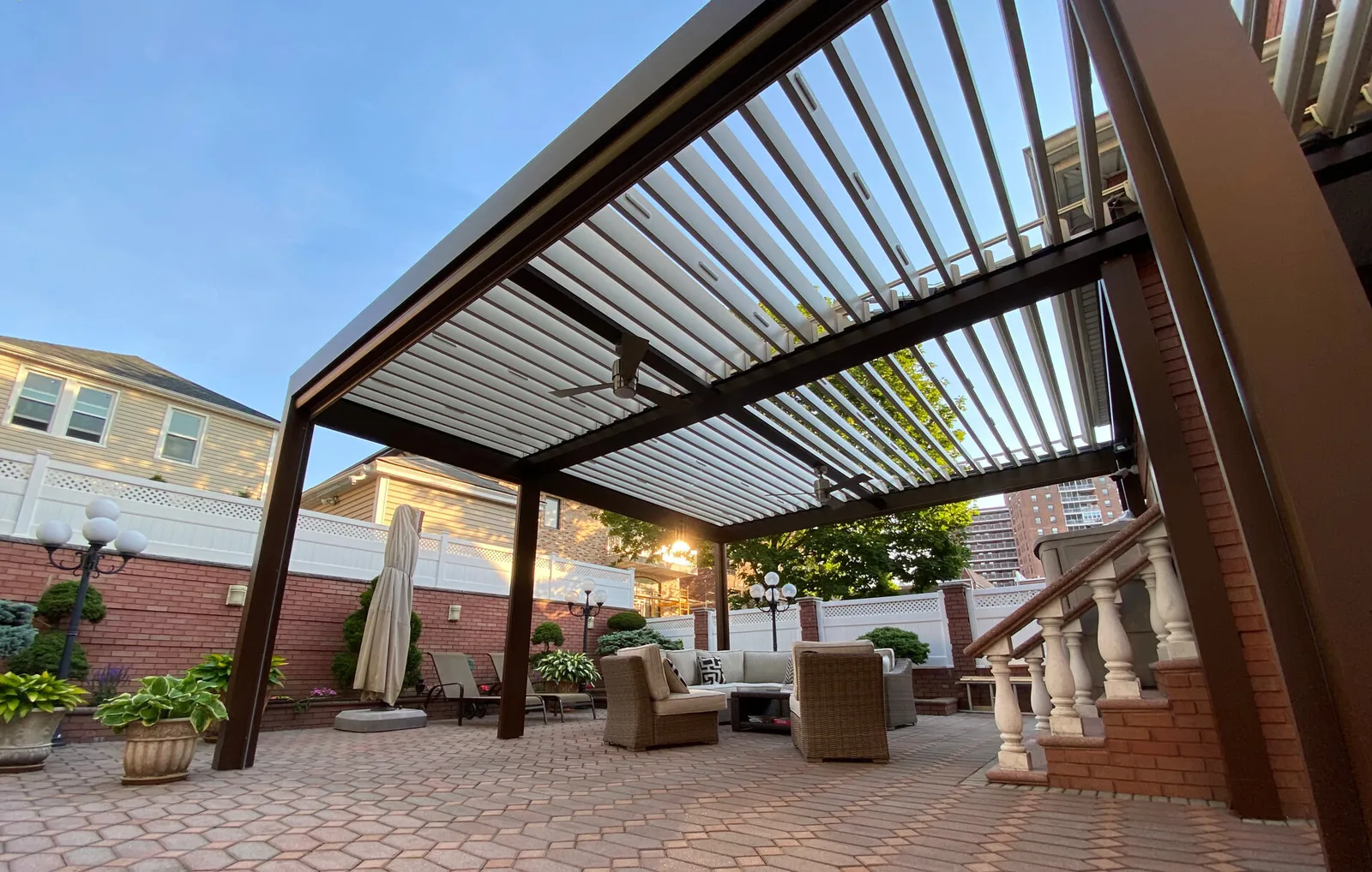 Bronze-framed louvered pergola in a brick courtyard at dusk, blades open with the sky showing through — photograph