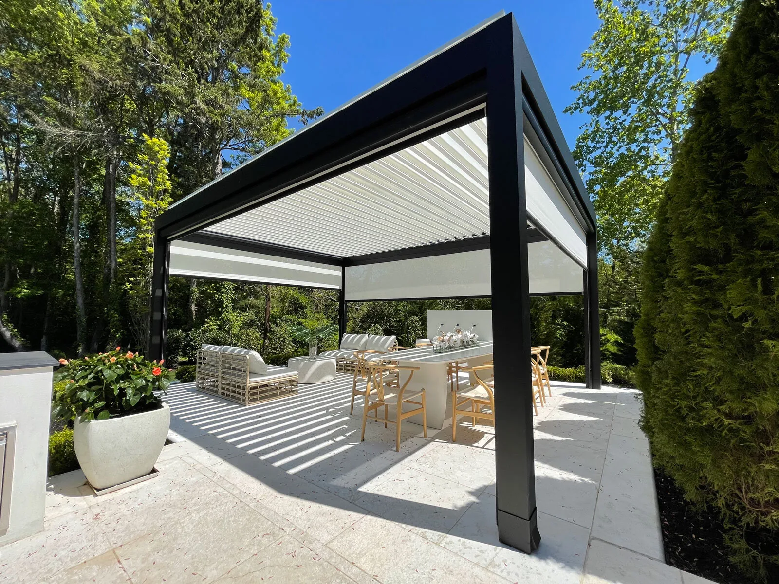 Black-framed louvered pergola with white blades angled over a stone terrace, striped shadows on the paving — photograph