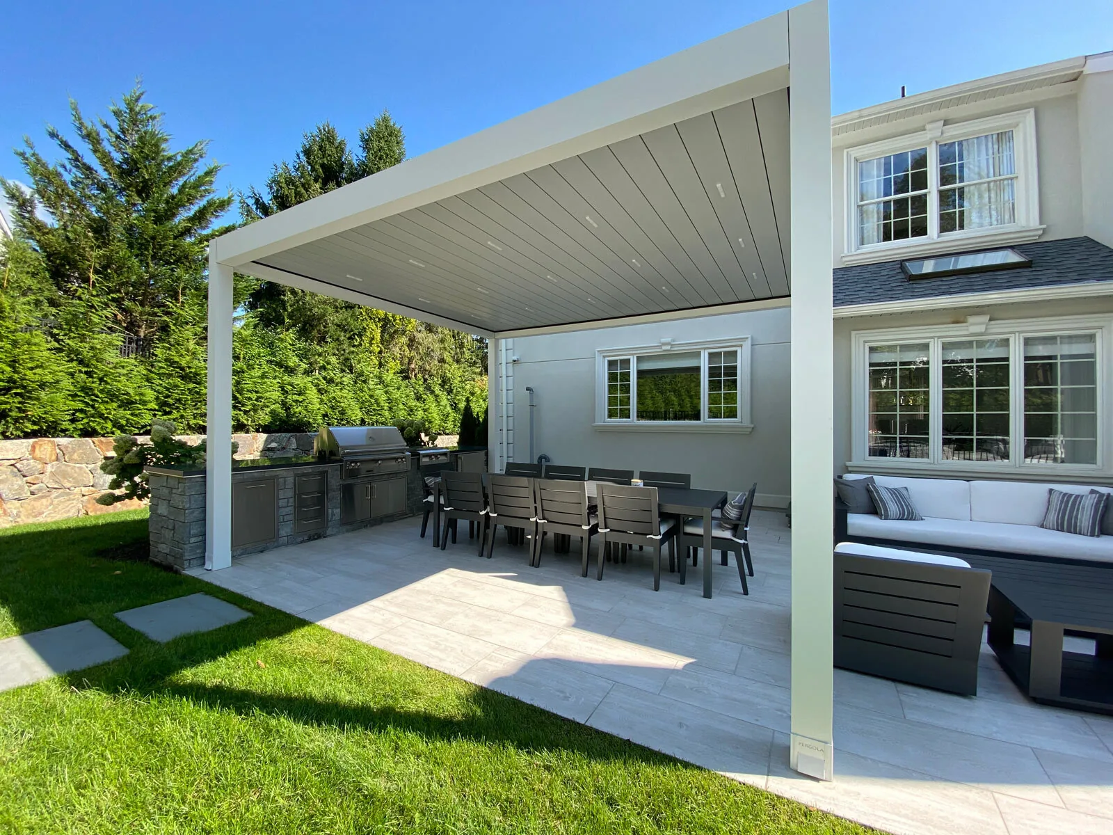 White-framed louvered pergola with the blades closed into a lit plane over an outdoor kitchen run — photograph