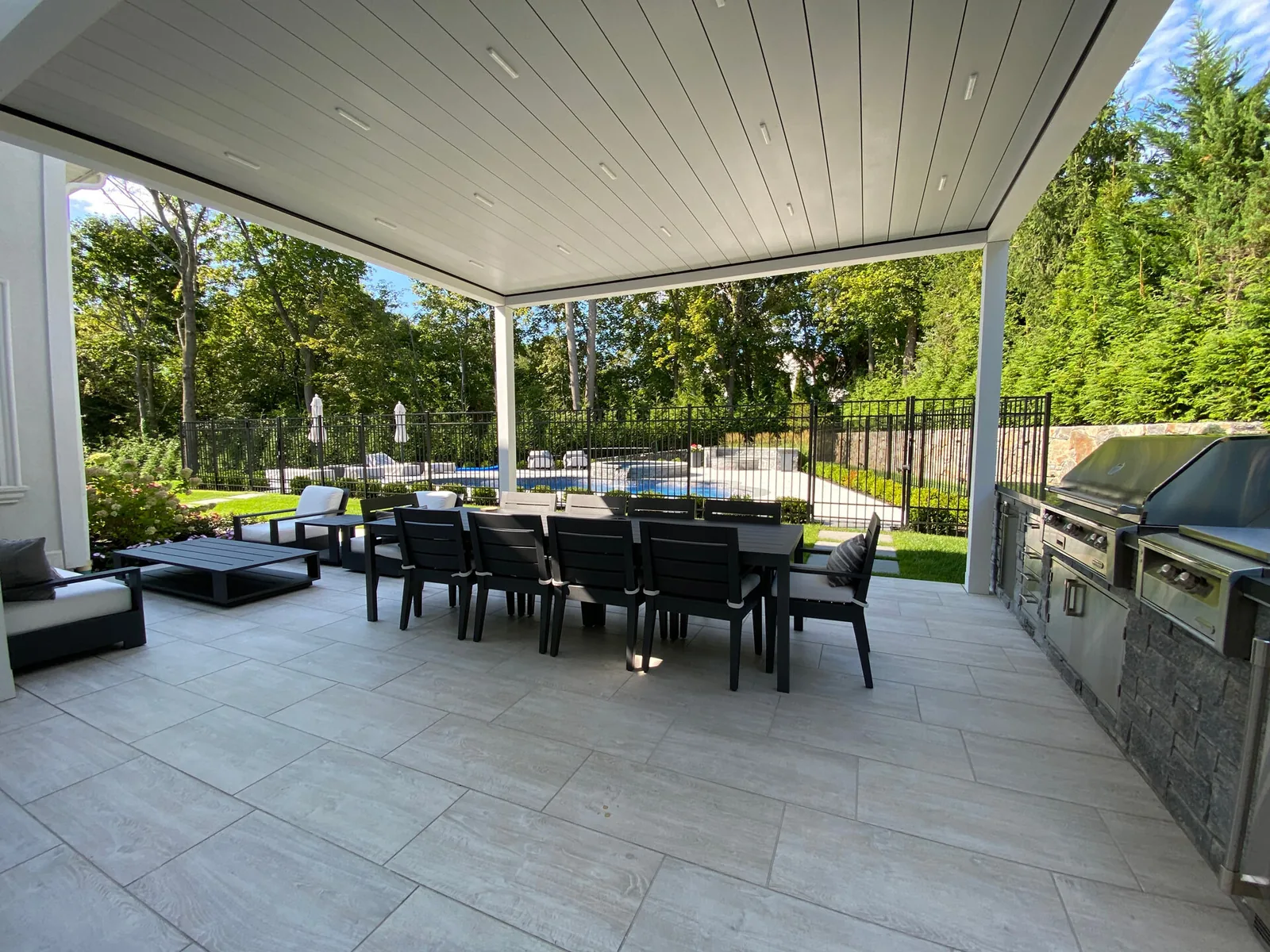 From under a white-framed louvered pergola with the blades closed and downlights set into the ceiling, dining table below and a pool beyond the fence — photograph