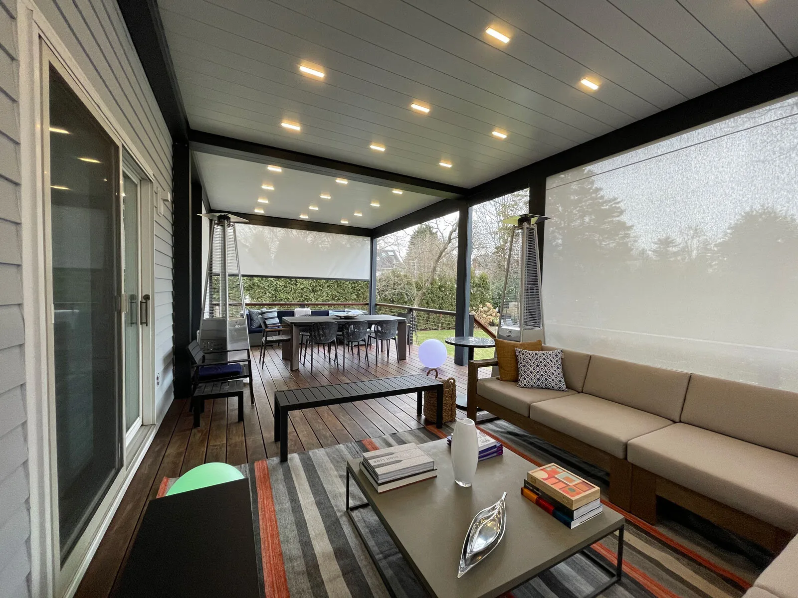 Inside a black-framed louvered roof on a wood deck, blades closed with downlights lit and screens down on the flank — photograph