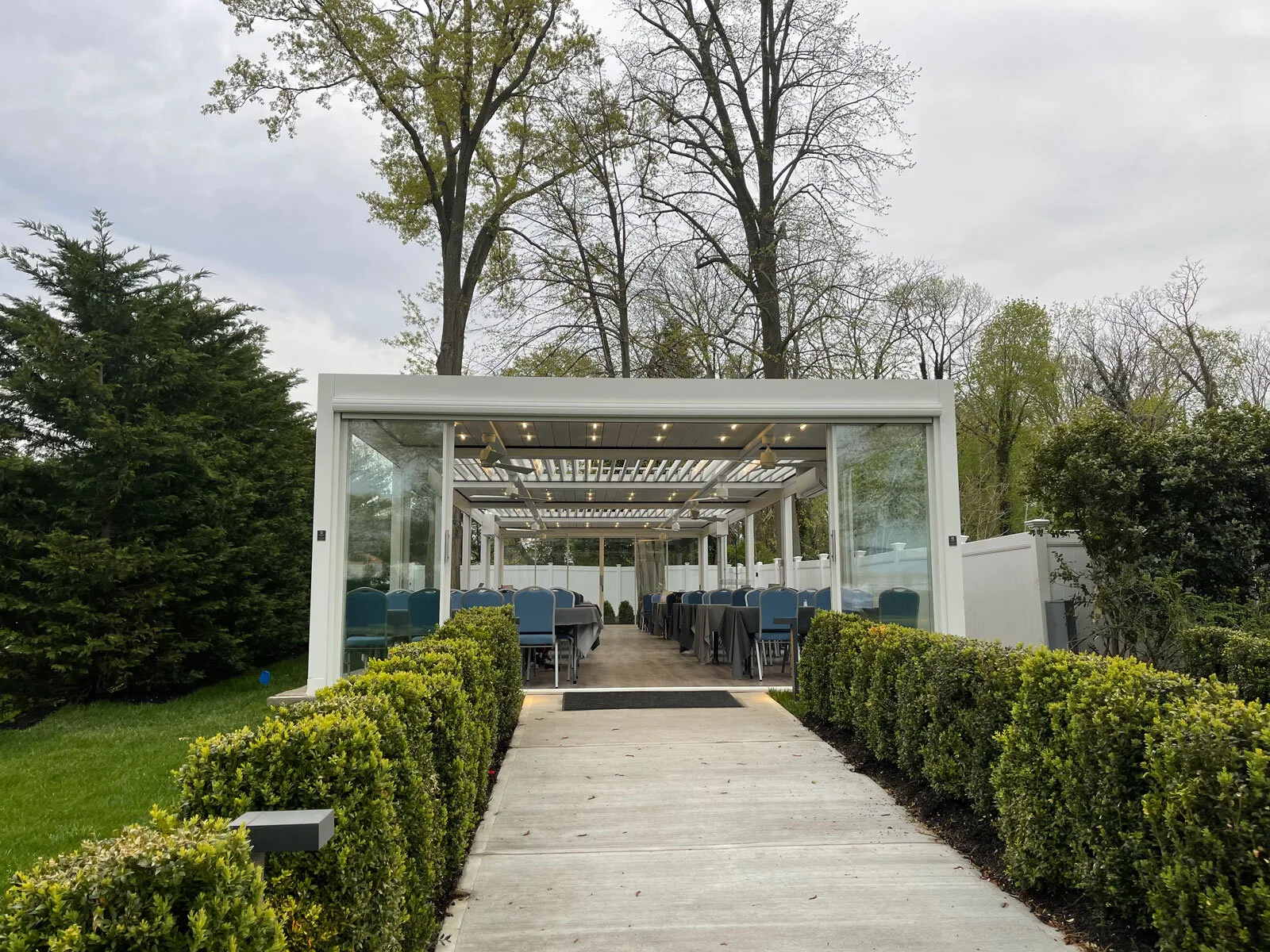 Freestanding white louvered pavilion enclosed with sliding glass on every side, banquet tables and chairs set inside — photograph