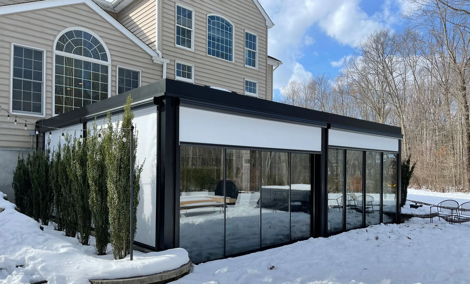 Black-framed louvered room attached to a house in snow, sliding glass walls below and zip screens lowered above — photograph