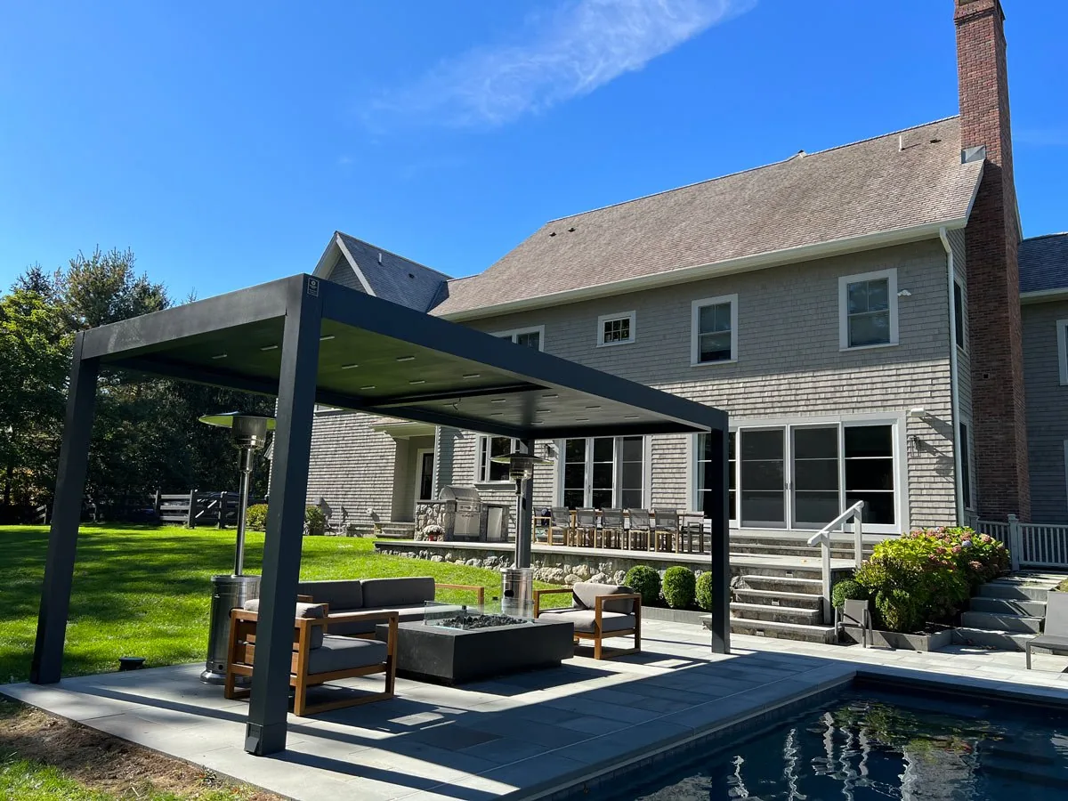 Freestanding charcoal louvered pergola beside a pool with the blades closed, fire table and teak seating beneath — photograph