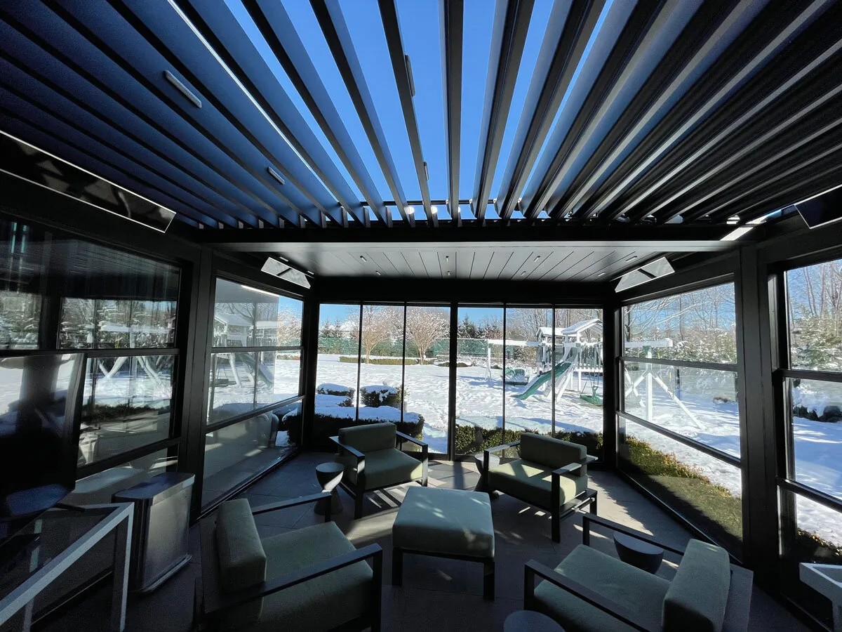 Inside a dark-framed room under a closed louvered roof, sliding glass walls shut against a snow-covered waterfront — photograph