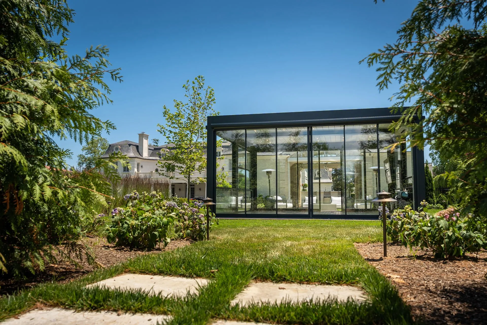 A dark-framed glass room on a lawn with its sliding glass wall closed along the long side, stepping stones leading to it — photograph