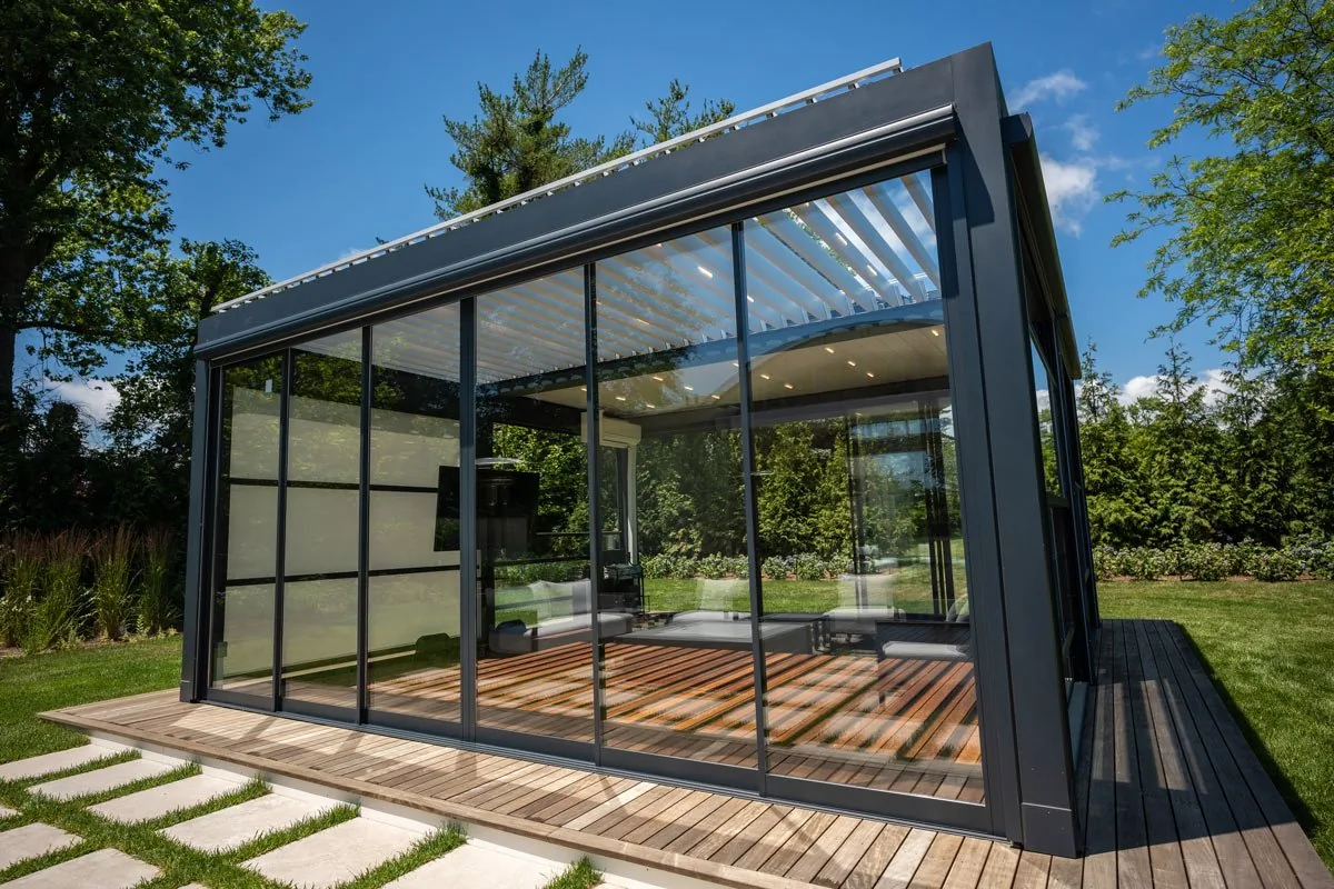 A dark-framed glass room on a wood deck with sliding glass panels closed, stepping stones across the grass to it — photograph