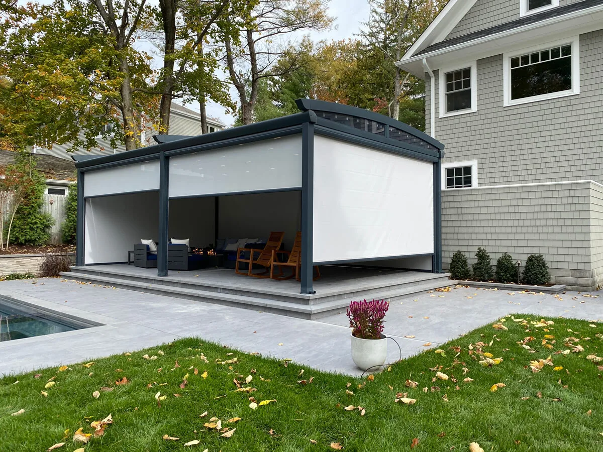 A dark-framed pergola beside a house with pale zip screens lowered along its long side — photograph