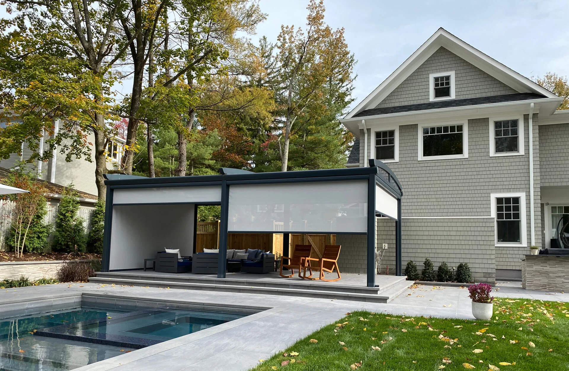 The same pergola seen across the pool, screens partly lowered, autumn trees behind the house — photograph