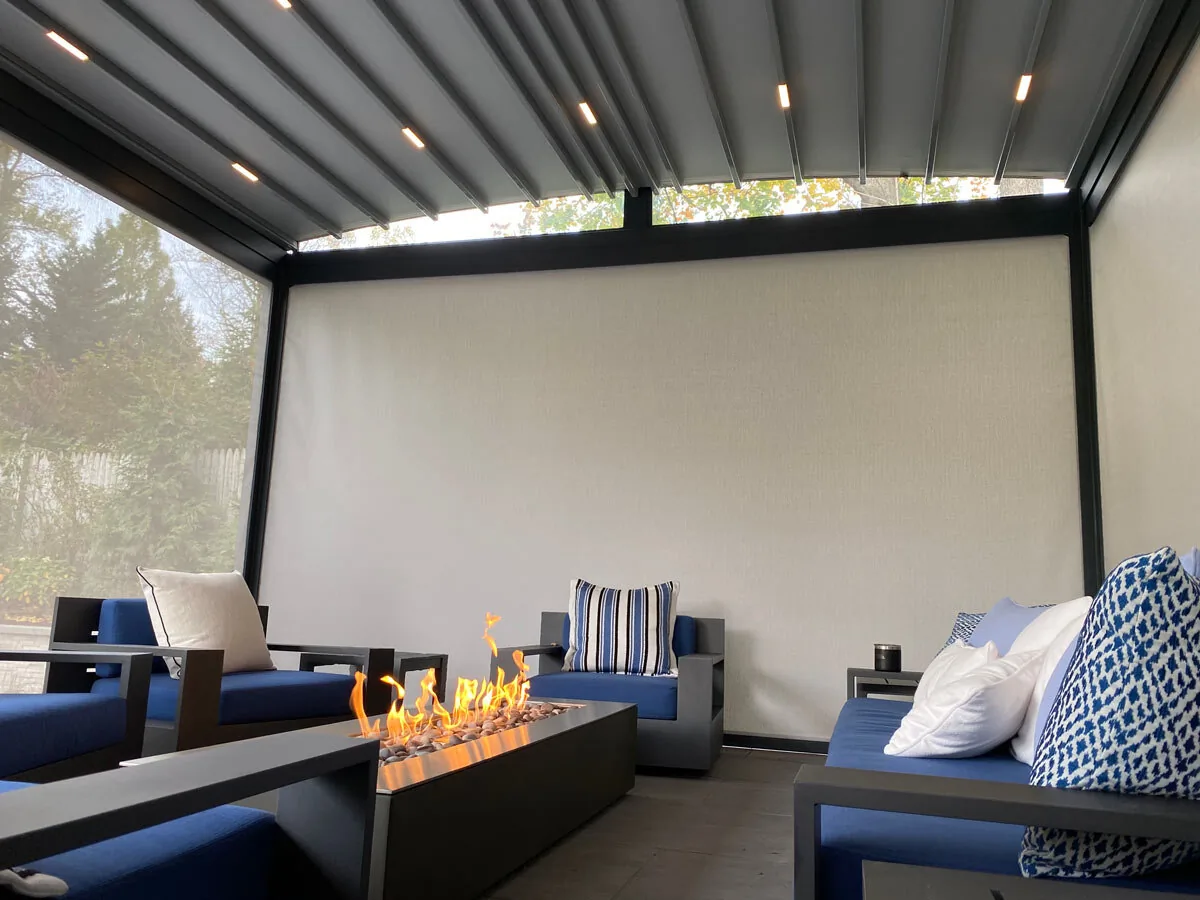 Inside the same pergola at night with the screens down, a lit fire table between blue cushioned seating under the closed blades — photograph